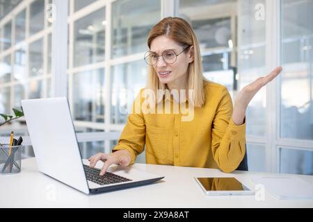 Confused professional woman with glasses using a laptop in a modern office setting, showing uncertainty and contemplation. Stock Photo