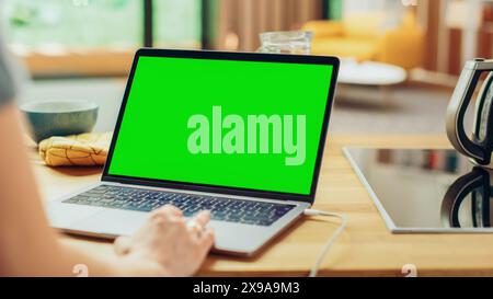 Close Up Green Screen Mock Up on a Laptop Computer. Device is Used on a Kitchen Table in a Modern Home. Person Uses Touch Pad for Scrolling the Web. Sunny Modern Kitchen with Healthy Lifestyle Vibes. Stock Photo