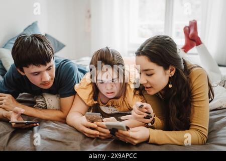 Curious male and female siblings sharing smart phone while lying down on bed at home Stock Photo