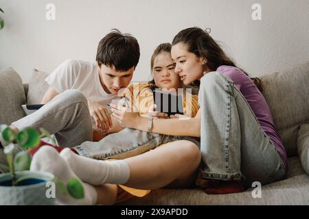 Curious male and female siblings sharing smart phone while sitting on sofa at home Stock Photo