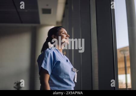 Smiling contemplative female doctor looking through window while standing at hospital Stock Photo