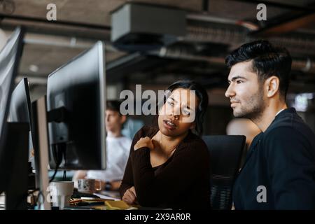 Young female programmer looking at monitor screen while sitting next to male colleague at office Stock Photo