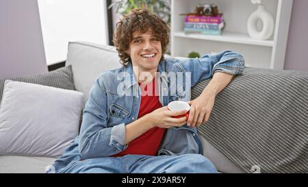 A smiling young man with curly hair relaxes on a sofa at home, holding a red mug. Stock Photo