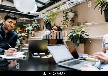 Smiling young female entrepreneur sitting with colleagues working on laptop at office Stock Photo