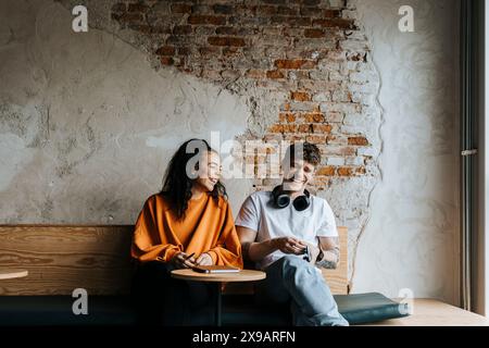 Happy male and female entrepreneurs laughing while sitting on bench against brick wall in office Stock Photo