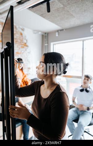 Young female entrepreneur writing on whiteboard in business meeting with colleagues at office Stock Photo