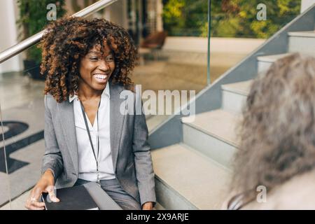 Happy female entrepreneur with curly hair sitting on steps while talking to delegate at convention center Stock Photo