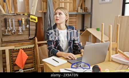 A blonde woman with blue eyes ponders a project in a cluttered carpentry workshop with tools and wood while working on a laptop. Stock Photo