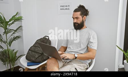 A young man with a beard works on a laptop in a modern waiting room with a 'free wi-fi available' sign. Stock Photo