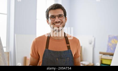 Smiling bearded man wearing glasses and an apron stands in a bright art studio. Stock Photo