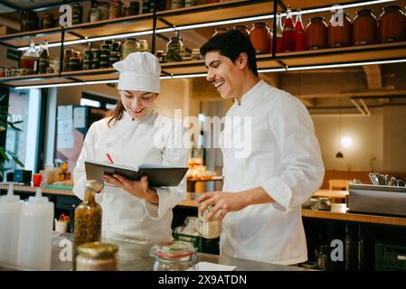 Happy female chef writing in diary with male coworker while discussing inventory in commercial kitchen Stock Photo