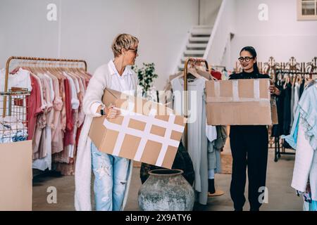 Female owner and colleague carrying cardboard boxes while working in clothing store Stock Photo