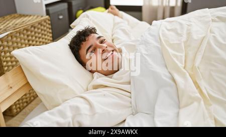 A cheerful young man with curly hair lying in bed, smiling in a bright bedroom setting. Stock Photo