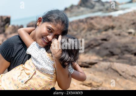 On a rugged coastline, a mother holds her child close, both gazing at the sea. Their bond is timeless, like the waves that break behind them. Stock Photo