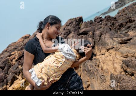 On a rugged coastline, a mother holds her child close, both gazing at the sea. Their bond is timeless, like the waves that break behind them. Stock Photo