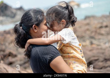 On a rugged coastline, a mother holds her child close, both gazing at the sea. Their bond is timeless, like the waves that break behind them Stock Photo