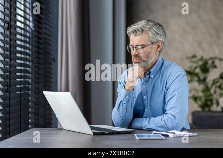 Senior man working on laptop in modern office environment, looking thoughtful. Natural light through window blinds creates a professional and focused atmosphere. Stock Photo