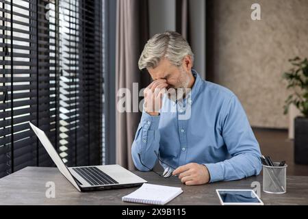 Senior businessman looking stressed while working on laptop at office desk. Depicts concepts of workplace stress, mental fatigue, and aging. Stock Photo