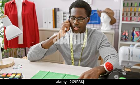 African american man in a tailor shop talking on the phone surrounded by sewing equipment and garments. Stock Photo