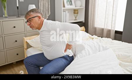 Mature man experiencing back pain while sitting in a modern bedroom interior. Stock Photo