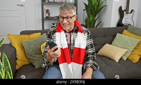 A cheerful, mature man interacts with a smartphone on a cozy sofa, surrounded by stylish home decor. Stock Photo