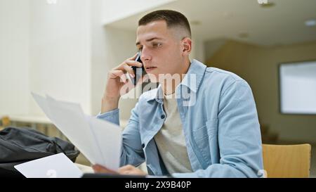 A focused young man uses a smartphone and reviews documents in a bright university library Stock Photo