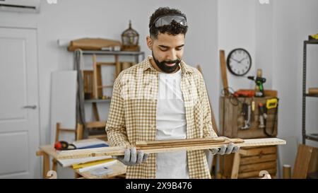 A young hispanic man examines a wooden plank with care in a well-equipped carpentry workshop Stock Photo