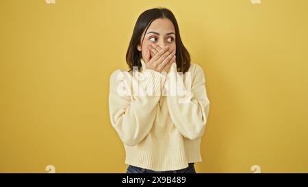 A young hispanic woman in casual attire gestures surprise against a vibrant yellow background. Stock Photo