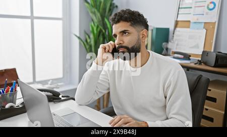 Handsome hispanic man with beard working on laptop in a modern office setting, looking pensive. Stock Photo