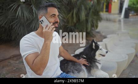 Handsome hispanic man talking on phone with husky dog in a sunny urban park. Stock Photo