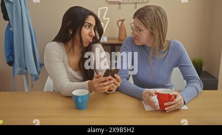 Two women friends share a humorous moment at a wooden table indoors, with cups and a smartphone. Stock Photo