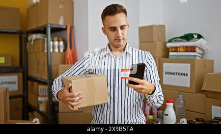 Handsome hispanic man checks smartphone while holding package in a donation center warehouse. Stock Photo