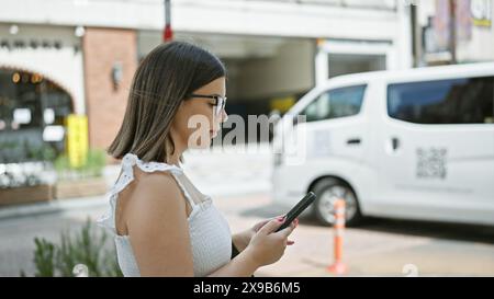 Beautiful hispanic woman with glasses walking confidently on tokyo streets while engrossed in her phone, typing a serious business text Stock Photo