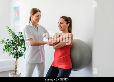 Physiotherapist helping woman do wall squats with fit ball. Young female patient doing back exercise using fitball in physio room of modern clinic Stock Photo