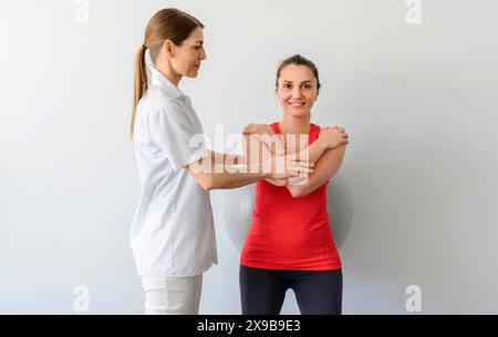 Physiotherapist helping woman do wall squats with fit ball. Young female patient doing back exercise using fitball in physio room of modern clinic Stock Photo