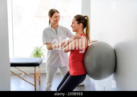 Physiotherapist helping woman do wall squats with fit ball. Young female patient doing back exercise using fitball in physio room of modern clinic Stock Photo
