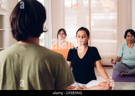 Group of people in a yoga class. Women and men practicing yoga on the purple mat in the lotus position with the trainer in front. Stock Photo