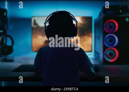 Low-light, high-contrast image of a boy playing a computer game on a gaming PC, viewed from behind, with a dramatic array of LED lights Stock Photo