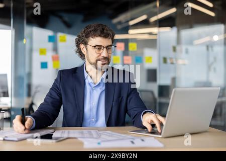 Serious focused businessman financier doing paperwork, accountant checking data from laptop and writing in paper tables, man in business suit experienced investor preparing report. Stock Photo