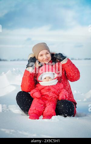 A smiling woman sits in the snow with a bundled-up baby, both dressed in warm winter clothing Stock Photo