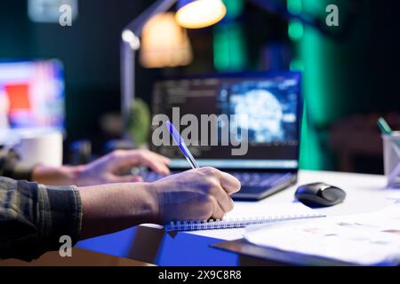 Male individual typing, and computing code for creating databases while writing on his notebook. Closeup of person working from home, monitoring software system and data center. Stock Photo