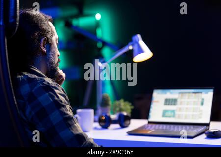 A bearded man checks economic and financial information on his portable computer. Male with hand on chin, looking at a laptop screen that displays the stock market. Stock Photo