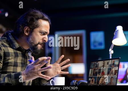 Frustrated male university student on a heated video conference with his colleagues. Angry man having a virtual meeting with group of coworkers, intense online discussion through laptop. Stock Photo