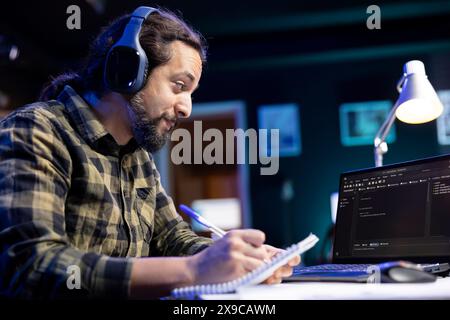 Man with notepad is working on laptop, programming code while listening to music on wireless headset. Male freelancer with headphones using monitoring system software on device and checking his notes. Stock Photo