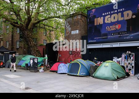 A person walks past a homeless encampment, seen with a row of tents under the backdrop of a noisy and busyTottenham Court Road. Stock Photo