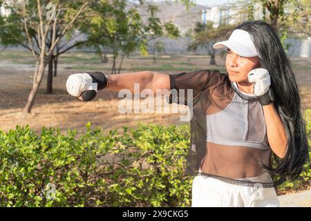 Woman in a transparent top and white cap practicing boxing in a park, focused on her punches under the sunny sky. Stock Photo
