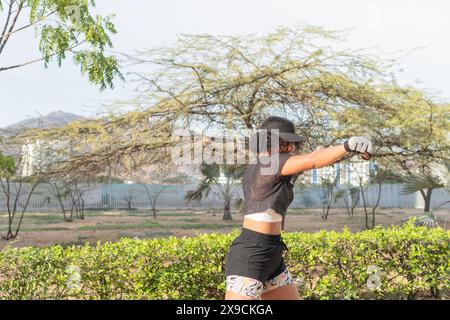 Woman in athletic wear and cap practicing boxing techniques outdoors in a sunny park, focusing on her stance and punches. Stock Photo