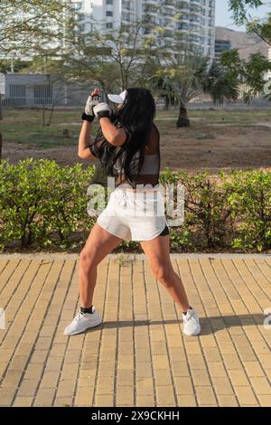Woman in a transparent top and white cap practicing boxing outdoors, with an urban backdrop, focusing on her movements in a sunny park. Stock Photo