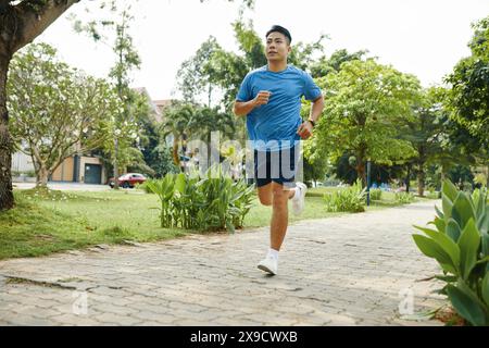 Young man in a blue shirt running along a park path during his morning exercise routine Stock Photo