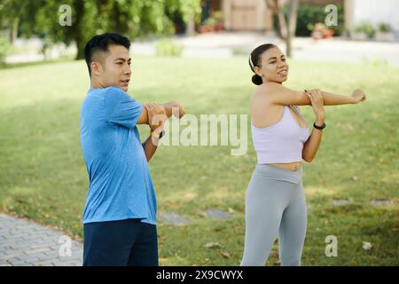 Man and woman in athletic wear stretching their arms together during an outdoor workout in the park Stock Photo